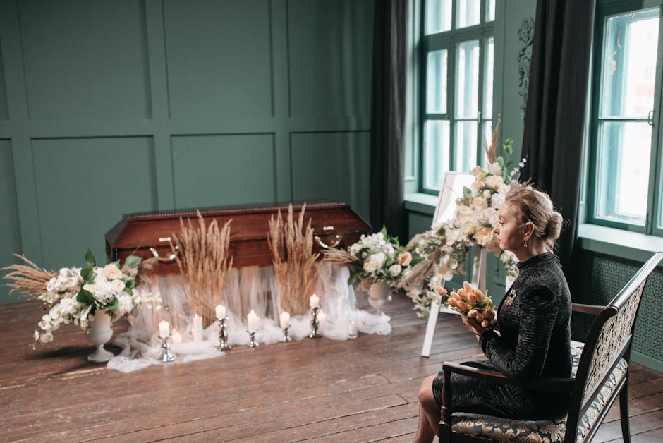 A woman seated in a dark, patterned dress holds a floral arrangement consisting of peach-colored roses and other soft-hued flowers. She is in a softly lit room with large, green-framed windows and dar