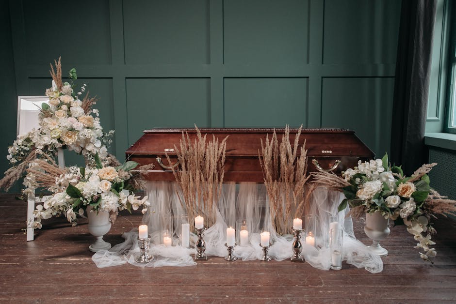 A solemn funeral arrangement featuring two large white floral bouquets placed on white pedestals, each containing cream roses, white hydrangeas, and greenery. In front of a polished wooden casket with