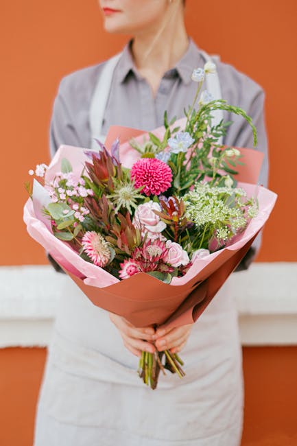 A florist from flowerswestheath.co.uk is holding a large, vibrant bouquet of fresh flowers, including pink and white roses, pink carnations, and agrestis-like greenery, all carefully arranged in pink 