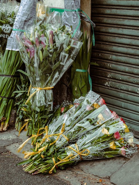 Multiple bouquets of fresh flowers, including pink lilies, yellow roses, and mixed greenery, wrapped in clear cellophane and tied with yellow ribbons, are arranged on the pavement outside a shop with 