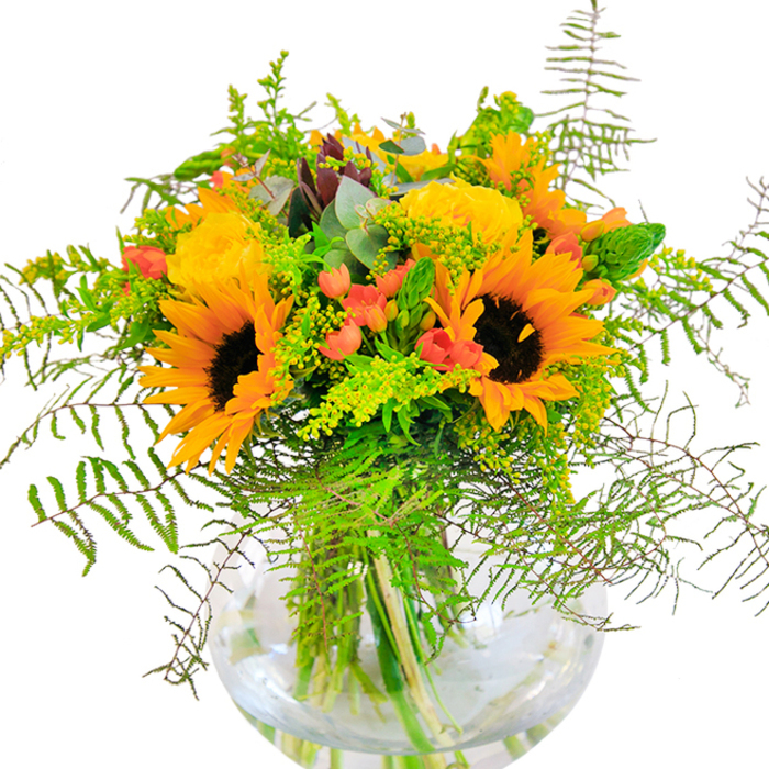 Upon first glance, this bouquet offers a radiant note of celebration that feels intrinsically tied to the spirit of West Heath. Towering sunflowers anchor the design, their dark, velvety centers framed by layers of brilliant yellow-orange petals that seem to bask in the soft daylight-a nod to the sunlit lawns of Golders Hill Park nearby. Classic garden roses, their petals unfurling in sumptuous spirals, bring richness and old-world charm. Contrasting with the bright blooms, clusters of red Hypericum berries glisten like tiny lanterns, their lively presence enlivening the verdant backdrop of cascading ferns, airy fillers, and arching stems. Wisps of eucalyptus, tinged blue-green, thread through the arrangement, evoking the wild, natural beauty of Hampstead Heath. Presented in a pristine, round glass vase, the bouquet's stems appear elegantly entangled, the fresh water glistening below in a dance of reflected hues. A gentle scent drifts upward-fresh, green, and inviting-while the overall composition radiates happiness. Handcrafted by Flowers West Heath, this arrangement would be equally at home at a birthday party, an anniversary dinner, or simply as an everyday delight to welcome friends and family to a home in NW3.
